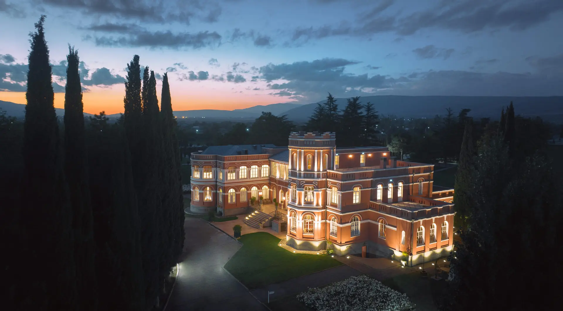 The palace of Château Mukhrani lit up at dusk among cypresses
