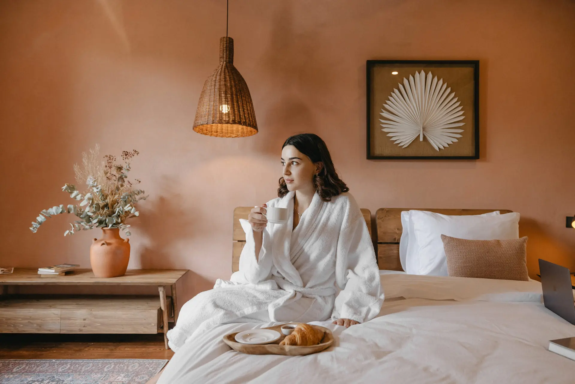 A guest with a cup of coffee on the bed of a terracotta room